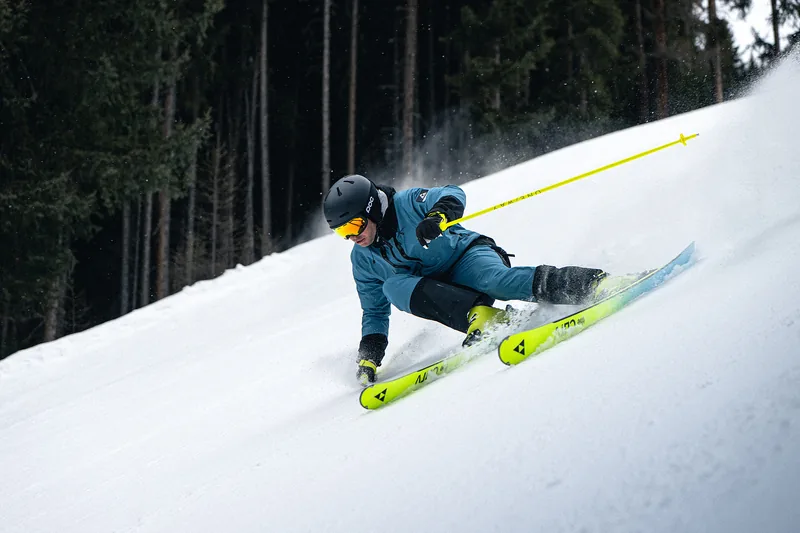 Skifahrer mit gelben Ski beim sportlichen Carven vor einer Waldkante.